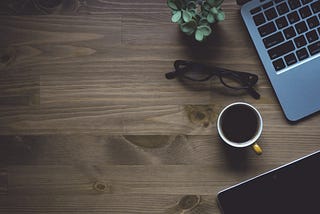 Laptop keyboard on a wooden desk next to a full coffee mug, reading glasses and a plant.