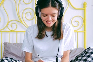 A young person sits on a bed. They are wearing headphones and using a tablet. The bed has a yellow frame against the wall.