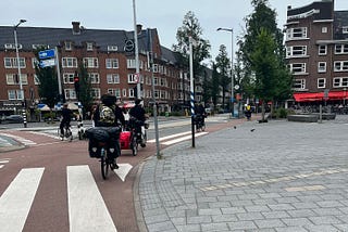Four bikes ride through the streets of Amsterdam. One has a dog trailer attached.