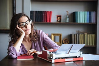 Dark haired woman stares pensively, her antique typewriter with sheet of paper sit on her desk.