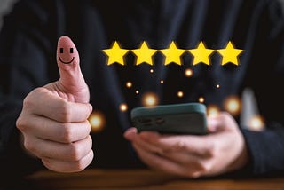 Five yellow stars in front of a person’s black-clad torso with one hand with a raised thumb with a smiley face on it and the other holding a phone.
