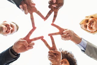 A diverse group of five people in professional attire are pictured from below as they hold out their fingers in V shapes and touch them together to form a star.