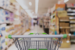 An image of a grocery cart standing in the middle of a grocery store aisle, from the perspective of someone pushing the grocery cart. The cart is in focus and the products on the shelves are blurred out.