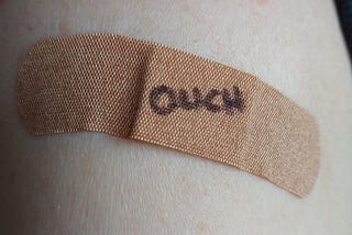 Photo of upper arm with generic bandaid, handwritten letters “OUCH” on bandaid