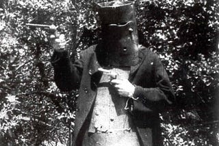 A black-and-white still showing an actor holding guns. His face is obscured by metal armor.