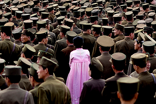 A woman stands in a crowd of North Korean soldiers wearing a pink Hanbok