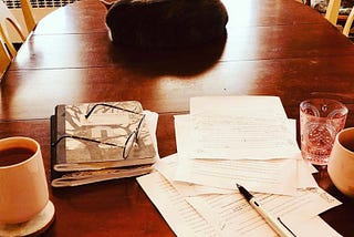A dining room table. In the foreground, two composition notebooks and several typewritten pages with notes scribbled across them, alongside a nearly empty glass of water and two mugs filled with hot beverages. Further back on the table, a cat. Beyond that, a trunk with some books on it. In the corner, a rocking chair.