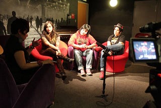 Dimly lit room with a spotlight on three bandmates in red chairs being interviewed.