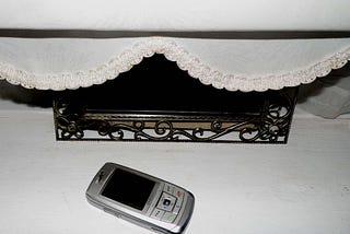 An old model of mobile phone on a white window ledge, in front of an ornately framed mirror, which is partly obscured by the scalloped edge of a roller blind.