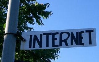 A plate in white at a mid length public light post which is written, in black, the word internet; a blue sky behind; tree top