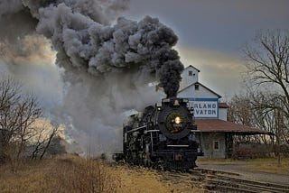 a train approaching on tracks with billowing black smoke rising up from the engine. The sky is ominous and dark and there are dead trees and dead yellow grass on either side of the tracks