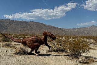 Rust colored herbivore dinosaur sculpture in the desert with gray mountains in background and blue sky with a few white clouds.
