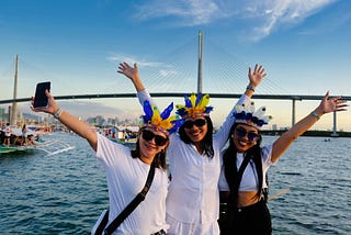Three smiling women wearing colorful feathered headpieces and sunglasses pose with their arms raised in celebration. They stand against a backdrop of a vibrant waterfront, with a modern suspension bridge and boats on the water during sunset. The scene reflects a festive and lively atmosphere