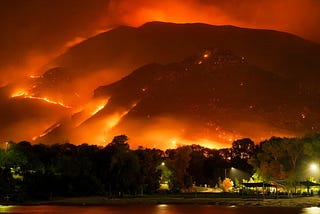 Rings of blazing fire engulf mountains  surrounding a town and reflect in the adjacent lake.