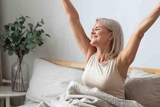 Middle agesd senior lady with white hair, white top, raising her arms in a stretch from a nap under white blanket and white pillows behind her. Teal coloured leaf arrangement on table on side of bed. She’s smiling and her eyes are closed.