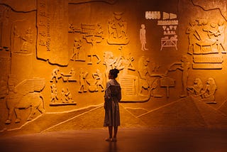 A person in trench coat standing in front of an ochre wall with huge hieroglyphics in relief