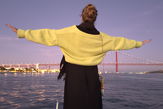 girl standing on the edge of a boat looking at the city of Lisbon in the background and having her hands in the air
