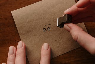 A hand holding a stamp, with the word No on brown kraft paper.