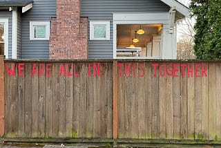 The words “We are all in the this together” are painted in large red letters on a brown fence in front of a grey house.