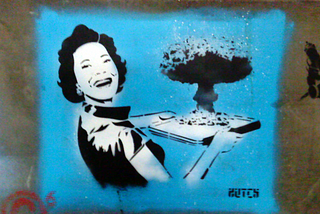 Photo of street art on a concrete wall. Against a blue background, a smiling 1950s housewife in black and white carries a serving tray containing a mushroom cloud. Via Wikimedia Commons/James Nash (aka Cirrus), license CC 2.0.