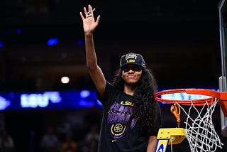 Angel Reese waving to the crowd after winning the NCAA Title. She is wearing a Black “National Champs” hat and shirt and is standing on a yellow ladder next to the basketball rim.
