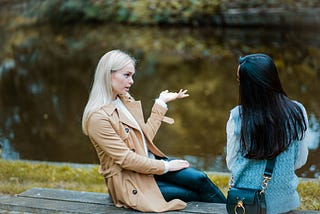 Two women sitting on a bench beside water, talking.