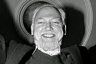 Uma foto preto e branco de Robert Anton Wilson, um homem branco, de origem irlandesa, um pouco careca, com um pouco de barba, pelos brancos, sorriso no rosto e roupas escuras.