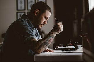 homem tatuado segurando um lapis, sentado numa cadeira em frente a um caderno branco