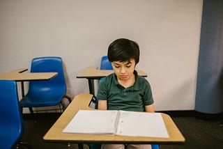 A young boy sits alone at a school desk, looking down at an open but empty notebook. He wears a dark green polo shirt and appears thoughtful or sad. Bright blue chairs and beige desks surround him in a plain classroom setting.