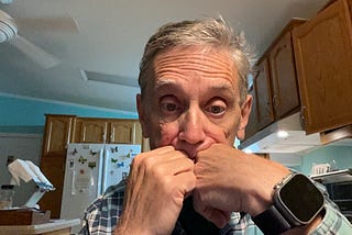 An older man sits in a kitchen, resting his face against both fists in a thoughtful or concerned pose. He is wearing a plaid shirt and an Apple Watch. The background includes wooden cabinets, a white refrigerator with butterfly magnets, and teal-colored walls.