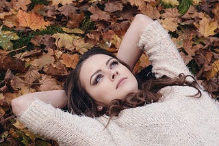 Young Attractive Brunette Woman In White Jumper Lying Down On Grass With Autumn Leaves.