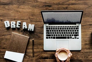 A person is holding a cup of coffee with both hands infront of a laptop. The word dream is pinned on the desk with a diary