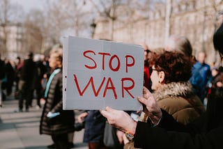 A person, whose face is out of frame, holds a signs that reads “STOP WAR” during a gathering of people outdoors. There are old neo-classical buildings and trees without leaves in the background.