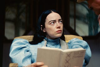 Bella Baxter (Emma Stone), a white woman with black hair and blue eyes, wears a Victorian gown and reads Love and Friendship.