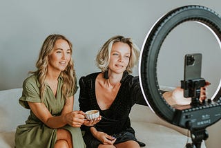 Two women sitting on a couch, one holding a cup, while the other adjusts a smartphone mounted on a ring light for filming or taking a photo. They appear relaxed and focused, possibly preparing for a video recording or livestream.