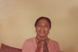 My grandmother sitting on a sofa, wearing a pink shirt and brown print scarf.