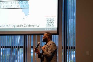 Dr. Jackie Beard in a gray blazer stands at the front of a conference room, holding a microphone and addressing attendees. Behind him, a large projection screen displays a presentation for the Association of Florida Colleges Region IV Conference, while floor-to-ceiling windows with vertical blinds line the room