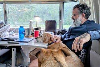 The author, a bearded man with glasses and a tattoo on his hand, is sitting inside a cozy RV. He is petting a light brown dog whose head is resting on the table. Another dog, also light brown, is sitting nearby with its back to the camera. The table holds various items including a laptop, a smartphone, a large blue water bottle, a red travel mug, and other personal items. Outside the window, it’s rainy with a view of lush green trees and a misty atmosphere, adding to the cozy indoor.