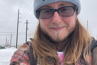 The author is wearing a knitted blue cap reading “Joy as Resistance”, gray sunglasses, a beard, long brown hair, a brownish coat, and a shirt the color of the trans flag (blue, white, and pink). He’s smiling slightly and has heavy bags under his eyes.
