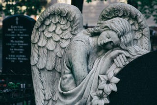 A stone statue of an angel leans peacefully against a graveyard headstone.