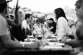 People sitting at a long dinner table outside, engaging each other in conversation