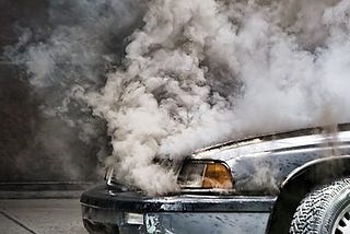 heavy, gray-black smoke roiling out from beneath the closed hood of a silver car