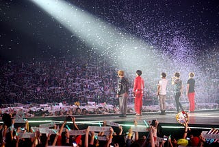 Five men stood on a stage in a spotlight in front of thousands of fans, all holding up banners.