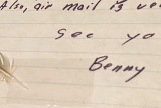 A portion of a handwritten letter on yellowing loose-leaf paper. There is a joke saying “Also, air mail is very expensive” followed by “See you soon! Benny.” A small yellow and dark gray feather has been taped to the left of the signature.