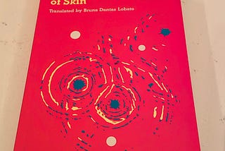 The cover of The Dark Side of Skin by Jeferson Tenorio is a vivid red with swirls of an abstract image in blue and yellow, perhaps making a female body. The book was translated by Bruna Dantas Lobato.