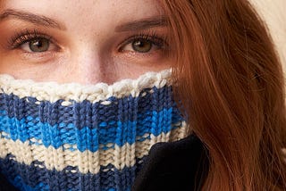 redheaded young woman has a turtleneck sweater pulled up over her mouth and nose, so that only her eyes are showing.