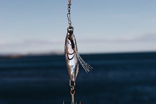 Silver fishing lure with many-pronged hook haning from a line with the ocean in the background.