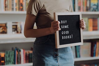 Person in brown t-shirt, standing in front of a bookshelf, holding a letter-board that says “BUY SOME GOOD BOOKS!”