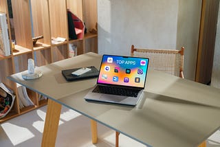 Image of a laptop on top of a wooden table with iPhone and iPhone and a candle to the left of the MacBook. Within the laptop’s screen is the text top apps with 10 app icons around it.