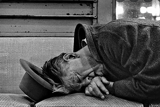 A black and white photo of a dirty man sleeping on a bench with his hat off near his head, circa 196o’s